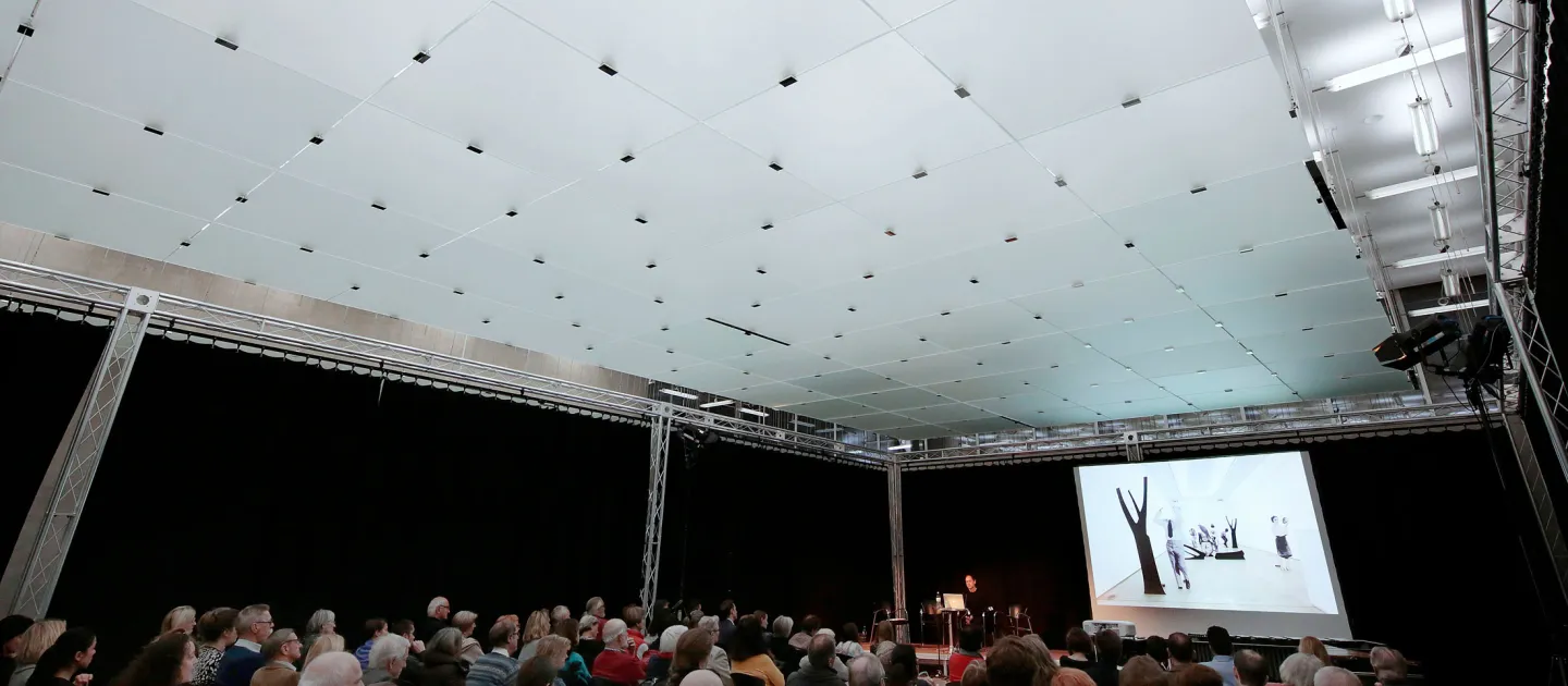 Szene aus "Einblick 2 – Gemeinsam zum Leuchtturm", 2015 im Kunsthaus Bregenz, Publikum sitzt auf Stühlen und blickt nach vorne auf eine Präsentation