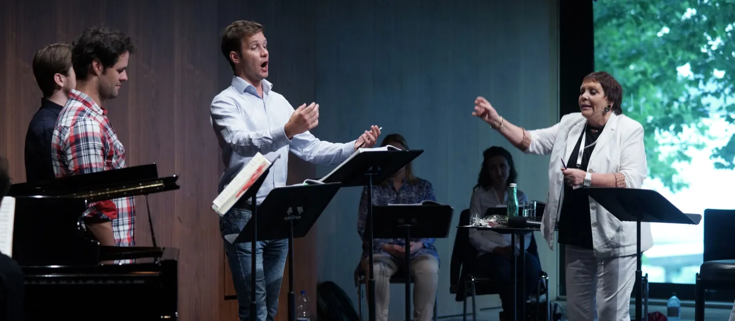 Szene aus "Meisterklasse Brigitte Fassbaender", 2015 im Festspielhaus, rechts Brigitte Fassbaender mit erhobenem Arm, drei weitere Personen stehen hinter einem Pult, zwei weitere Personen sitzen im Hintergrund hinter einem Pult