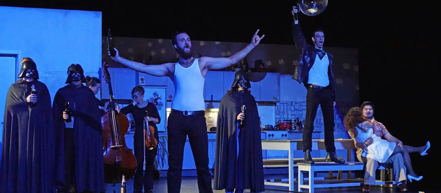Szene aus "Don Giovanni", 2016 im Theater am Kornmarkt, mehrere kostümierte Darstellende, zentral eine Person in weißem Tanktop mit ausgestreckten Armen, im Hintergrund drei Personen mit Darth-Vader-Maske und schwarzem Umhang, zwei Personen, eine mit einem Kontrabass, die andere mit Geige, rechts steht eine Person auf einer Bank und hält eine Diskokugel, zwei Personen sitzen auf einem Stuhl