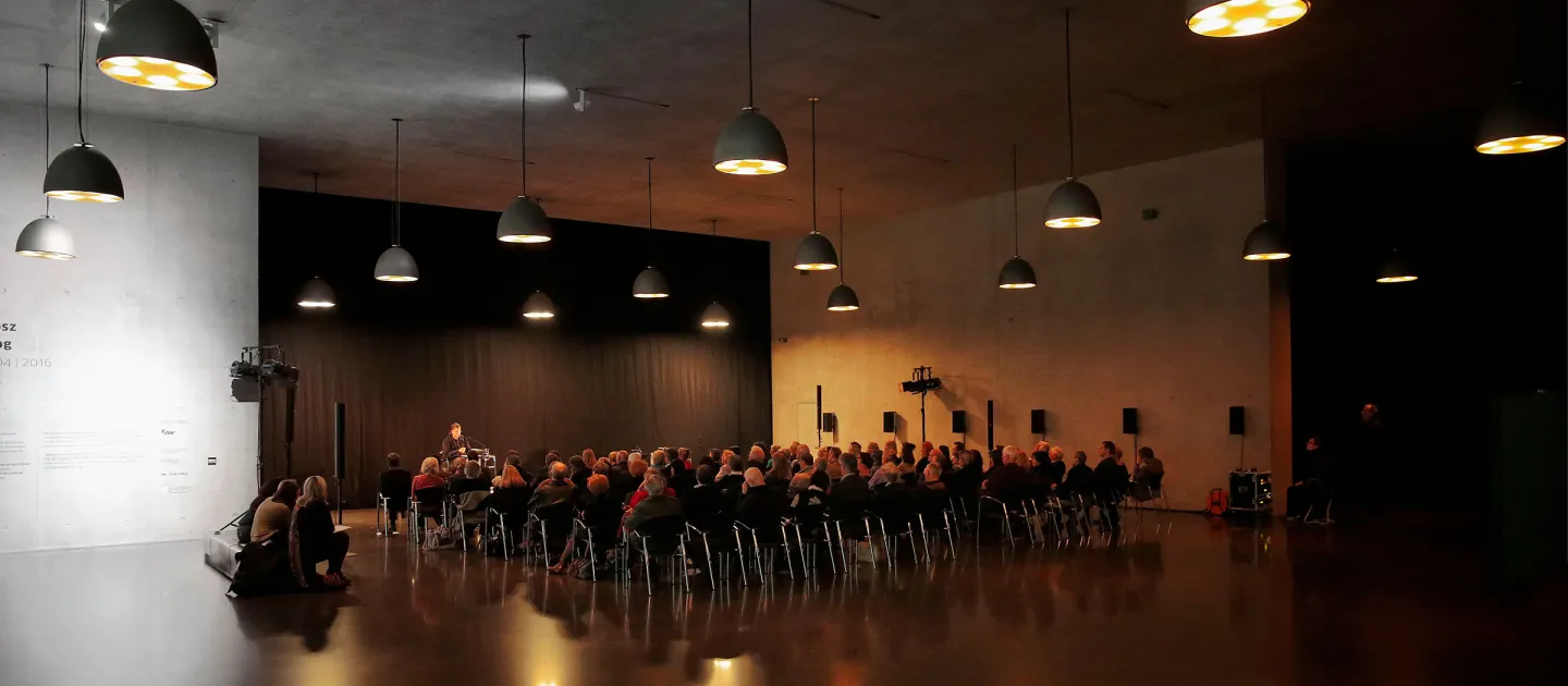 Szene aus "Einblick 3 - Gemeinsam zum Leuchtturm", 2016 im Kunsthaus Bregenz, Publikum sitzt in einem dunklen Raum, mehrere Lampen hängen von der Decke, vor dem Publikum befindet sich eine Person