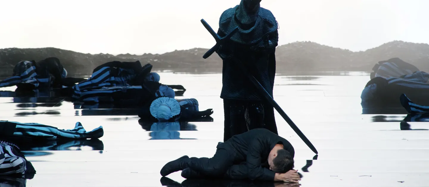 Szene aus "Hamlet", 2016 im Festspielhaus, Ritter in Rüstung mit langem Schwert, davor ein Mann am Boden liegend, reflektierender Boden, im Hintergrund weitere Personen am Boden liegend