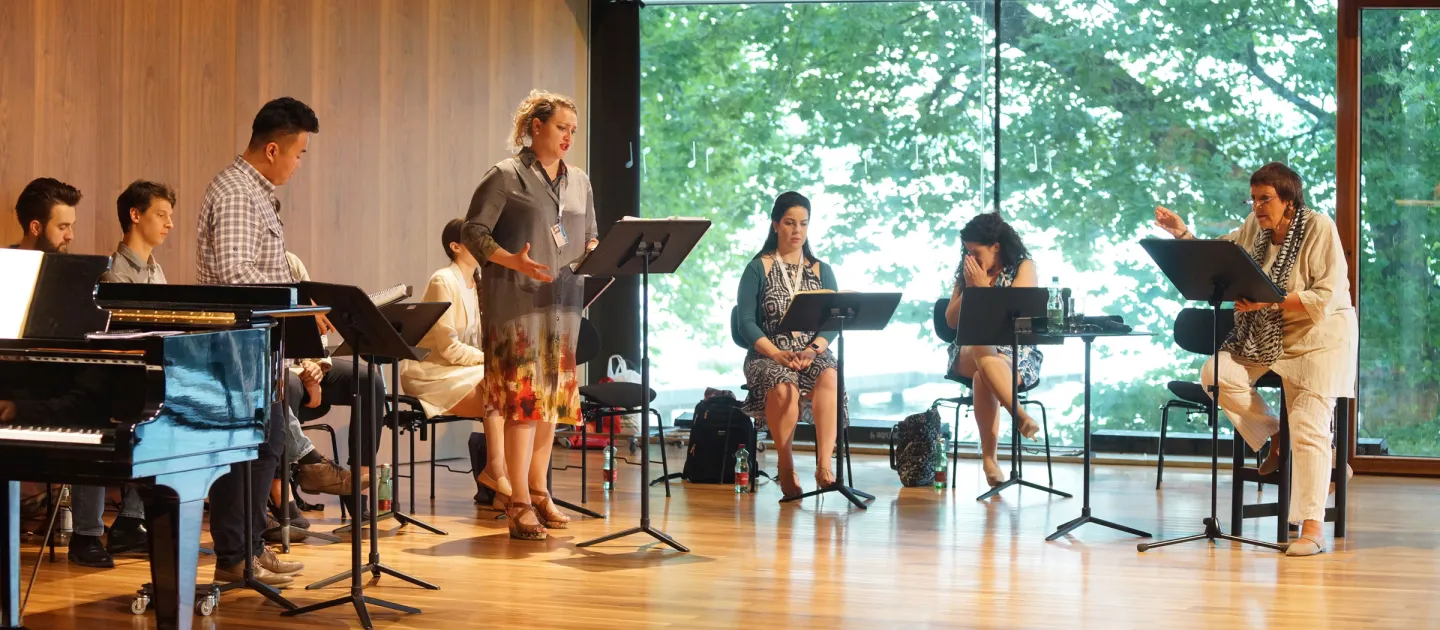Szene aus "Meisterklasse Brigitte Fassbaender", 2016 im Festspielhaus, mehrere Personen sitzen oder stehen hinter dem Pult, Brigitte Fassbaender rechts hinter dem Pult