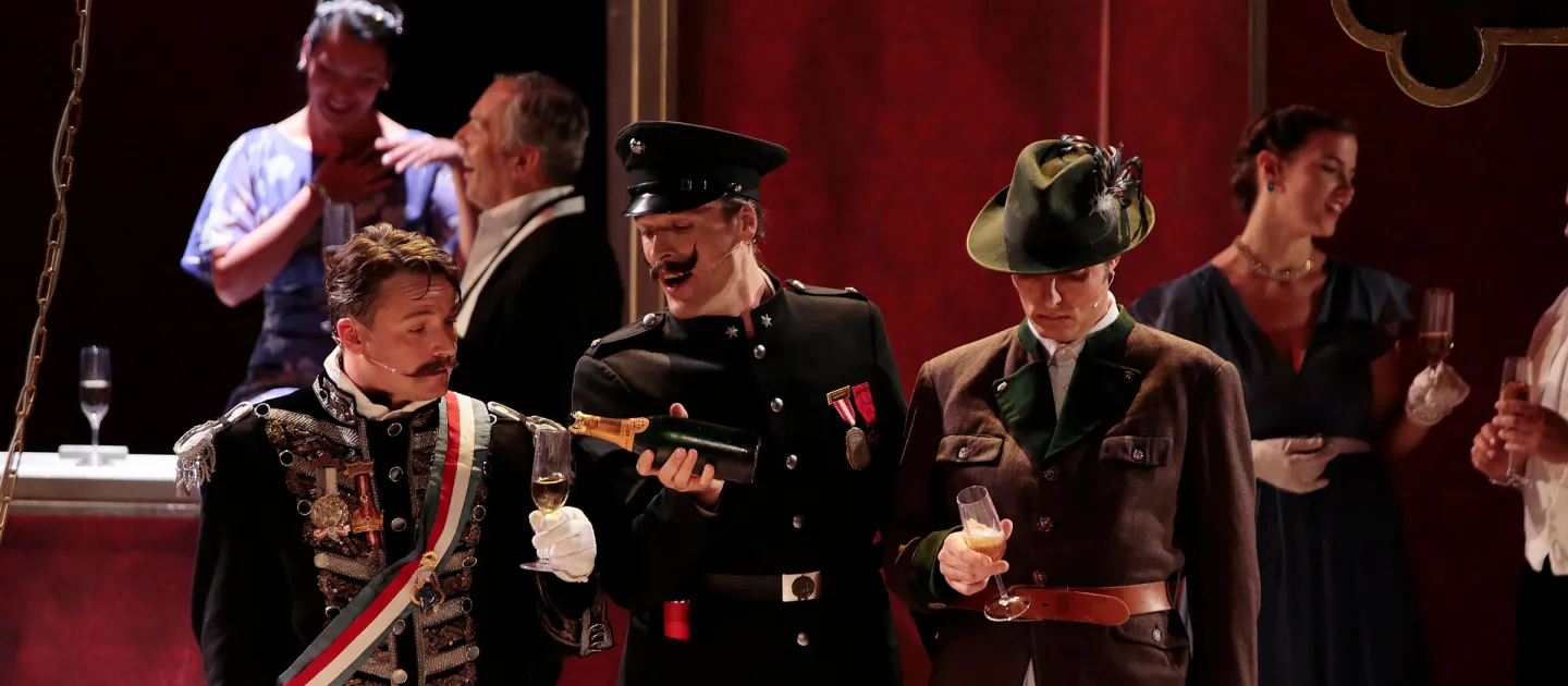 Szene aus "Staatsoperette", 2016 im Festspielhaus, im Vordergrund drei Darsteller in historischen Kostümen, zwei halten Sektgläser, im Hintergrund weitere kostümierte Personen