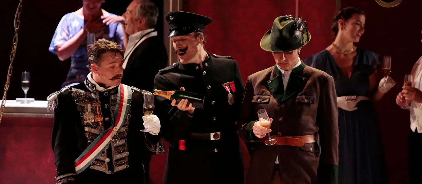Szene aus "Staatsoperette", 2016 im Festspielhaus, im Vordergrund drei Darsteller in historischen Kostümen, zwei halten Sektgläser, im Hintergrund weitere kostümierte Personen