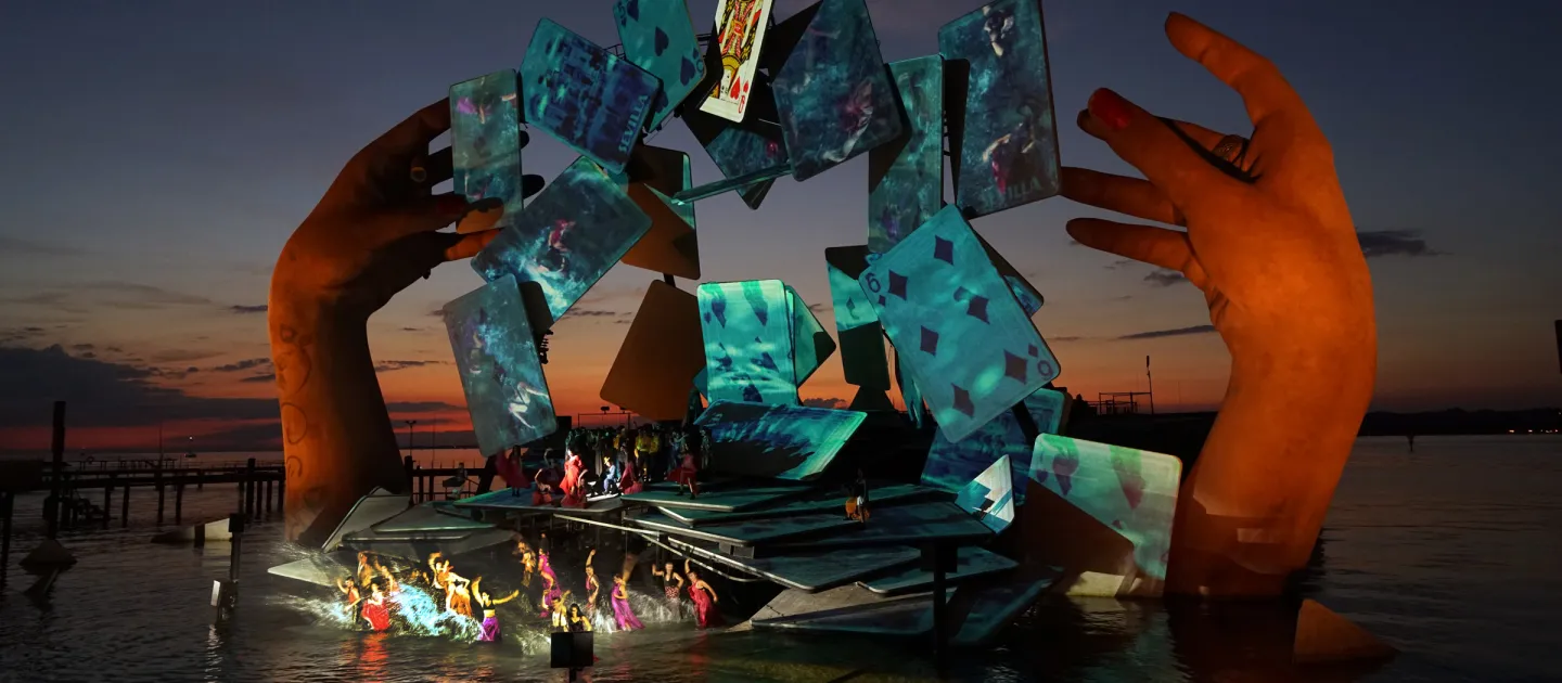Szene aus "Carmen", 2018 auf der Seebühne, große Hände über dem Wasser, schwebende grün-beleuchtete Spielkarten im Bogen, kostümierte Darstellende im Vordergrund auf der Bühnenfläche