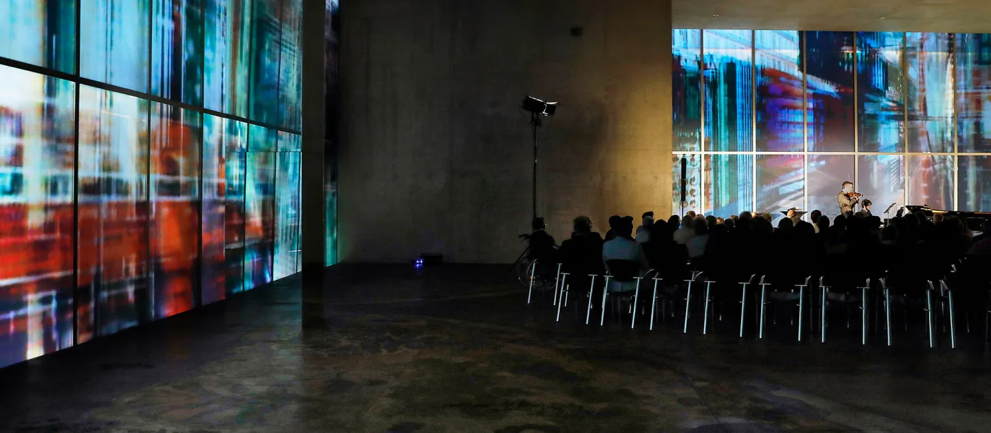 Szene aus "Konzert im KUB", 2017 im Kunsthaus Bregenz, Musiker:in auf der Bühne, davor Publikum auf Stühlen sitzend, Publikum von hinten im Weitwinkel fotografiert