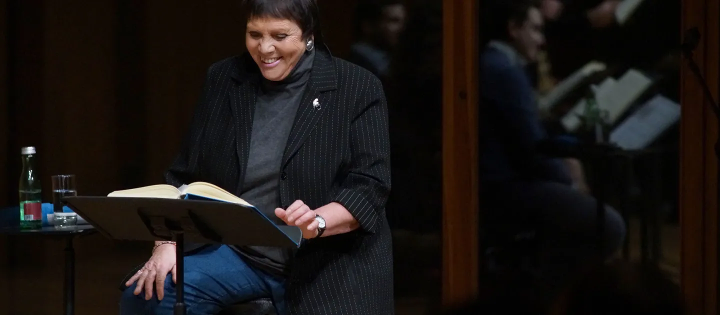 Szene aus "Meisterklasse B. Fassbaender", 2017 im Festspielhaus, B. Fassbaender sitzt lächelnd auf einem Stuhl, vor ihr ein Pult