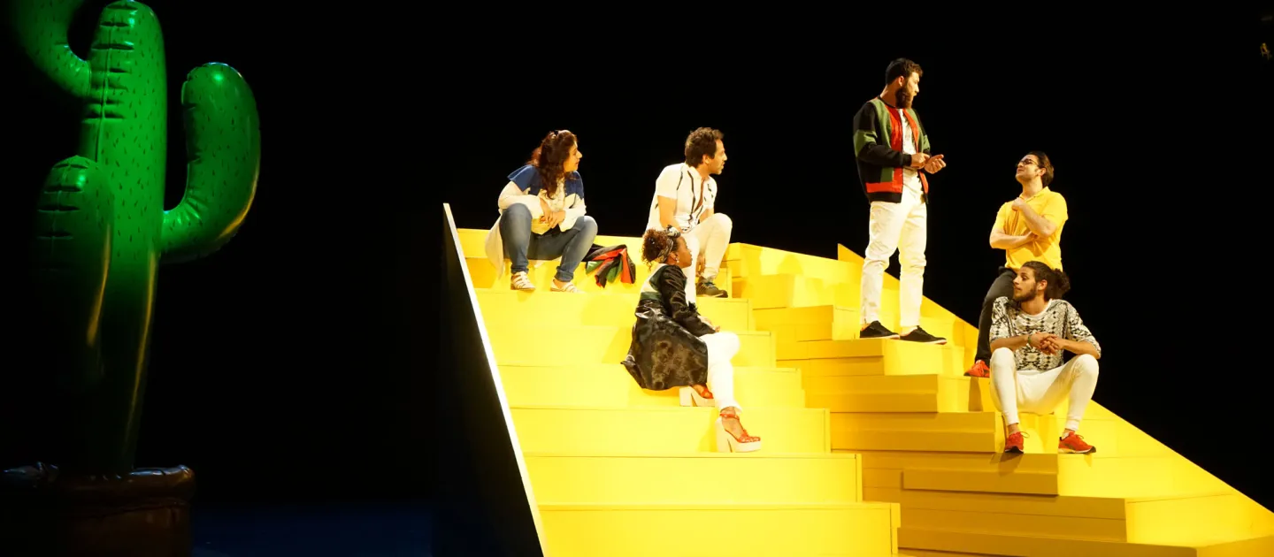 Szene aus "The Situation", 2017 im Theater am Kornmarkt, sechs kostümierte Darstellende auf einer großen gelben Treppe, zwei von ihnen stehen, links ein großer Luftballon in Kaktusform