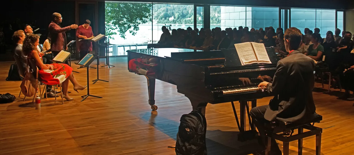 Szene aus "Meisterklasse Brigitte Fassbaender", 2018 im Festspielhaus, Flügel mit Pianist, vier Sänger:innen, davon eine Person stehend, Brigitte Fassbaender am Pult stehend, Publikum schaut zu und hört zu