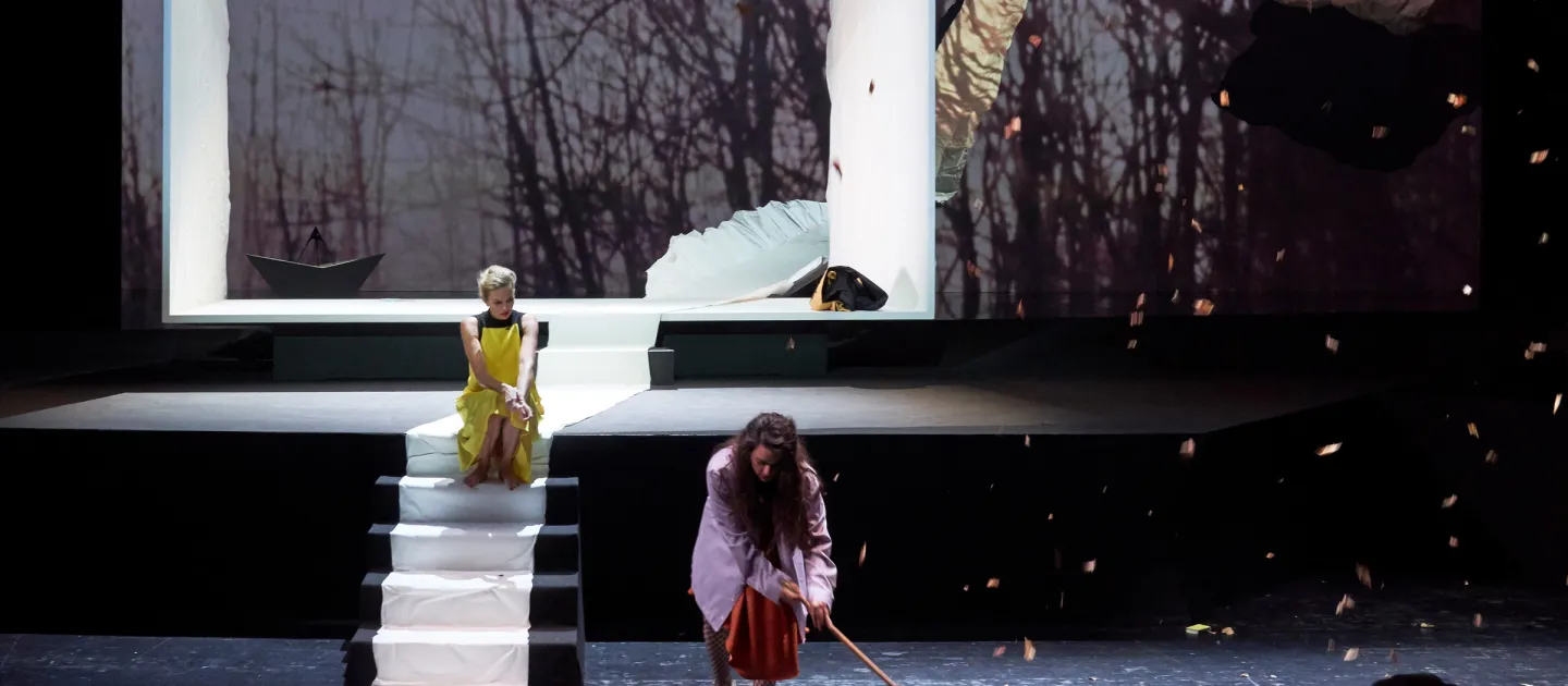 Szene aus "Das Jagdgewehr", 2018 im Festspielhaus, zwei Personen, eine sitzt auf weißen Stufen in gelbem Kleid, die andere kehrt mit Besen auf der Bühne, im im Hintergrund ein übergroßer weißer Rahmen und eine Projektion eines Waldes