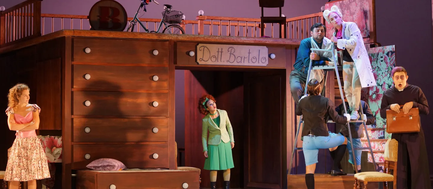 Szene aus "Der Barbier von Sevilla", 2018 im Theater am Kornmarkt, bunt kostümierte Darstellende auf der Bühne, zwei auf einem Hochstuhl, großes Holzmöbelstück mit Schubläden im Hintergrund