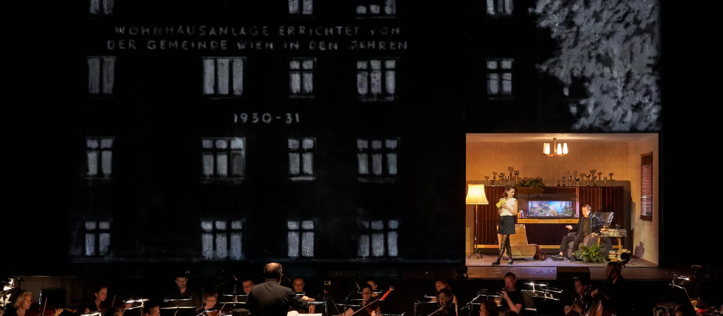 Szene aus "Der Reigen", 2019 im Festspielhaus, Orchester spielt vor Kulisse einer Wohnhausanlage mit Aufschrift "1930-1931", rechts Szene mit zwei kostümierten Darstellenden in Wohnzimmerkulisse