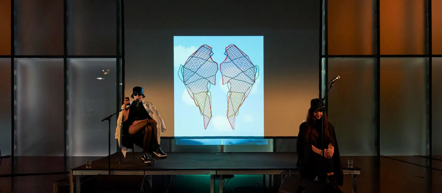 Szene aus "Einblick 2: Die Künstlerin Flaka Haliti", 2019 im Kunsthaus Bregenz, zwei kostümierte Darstellende auf der Bühne, im Hintergrund eine Projektion von zwei stilisierten Flügeln vor blauem Himmel mit Wolken