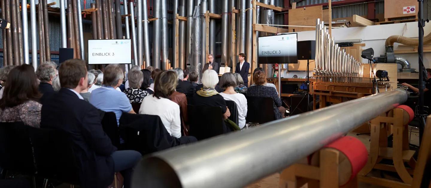 Szene aus "Einblick 3: Das Instrument", 2019 bei Rieger-Orgelbau Schwarzach, Publikum hört zwei Personen zu, umgeben Orgelpfeifen