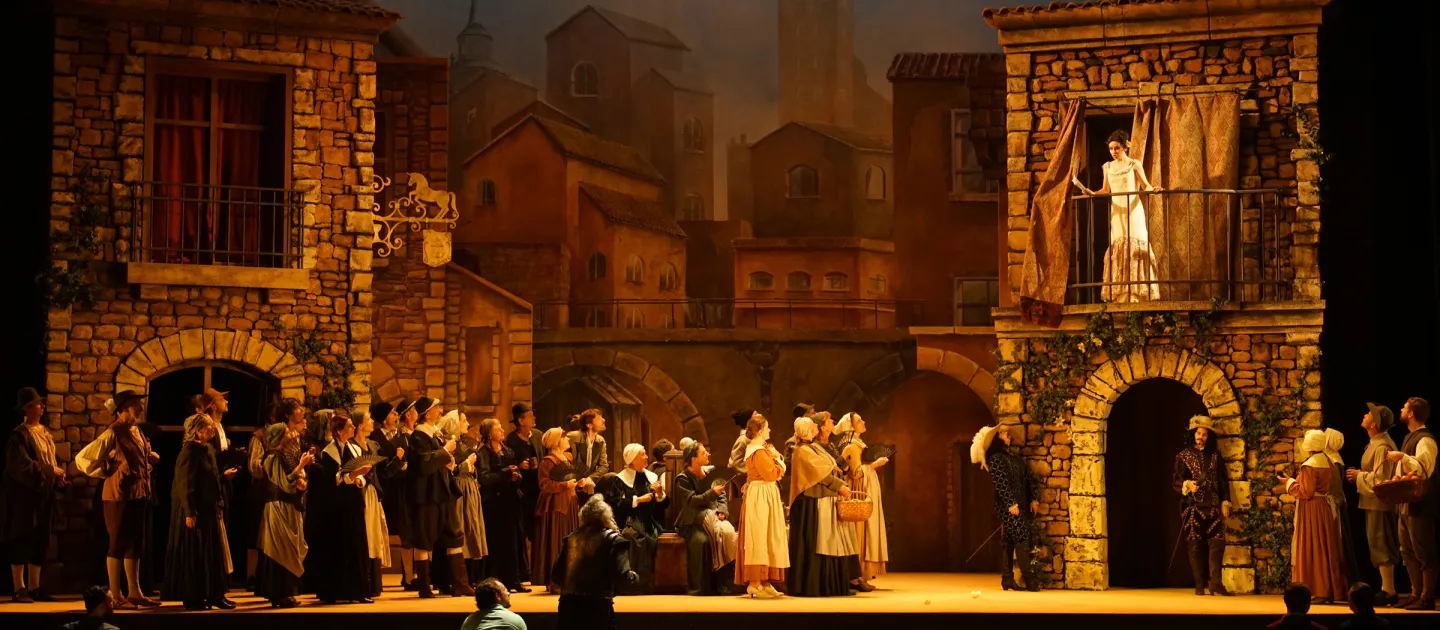 Szene aus "Don Quichotte", 2019 im Festspielhaus, viele kostümierte Darstellende vor Steinhaus-Kulisse, Darstellerin im weißen Kleid auf dem Balkon