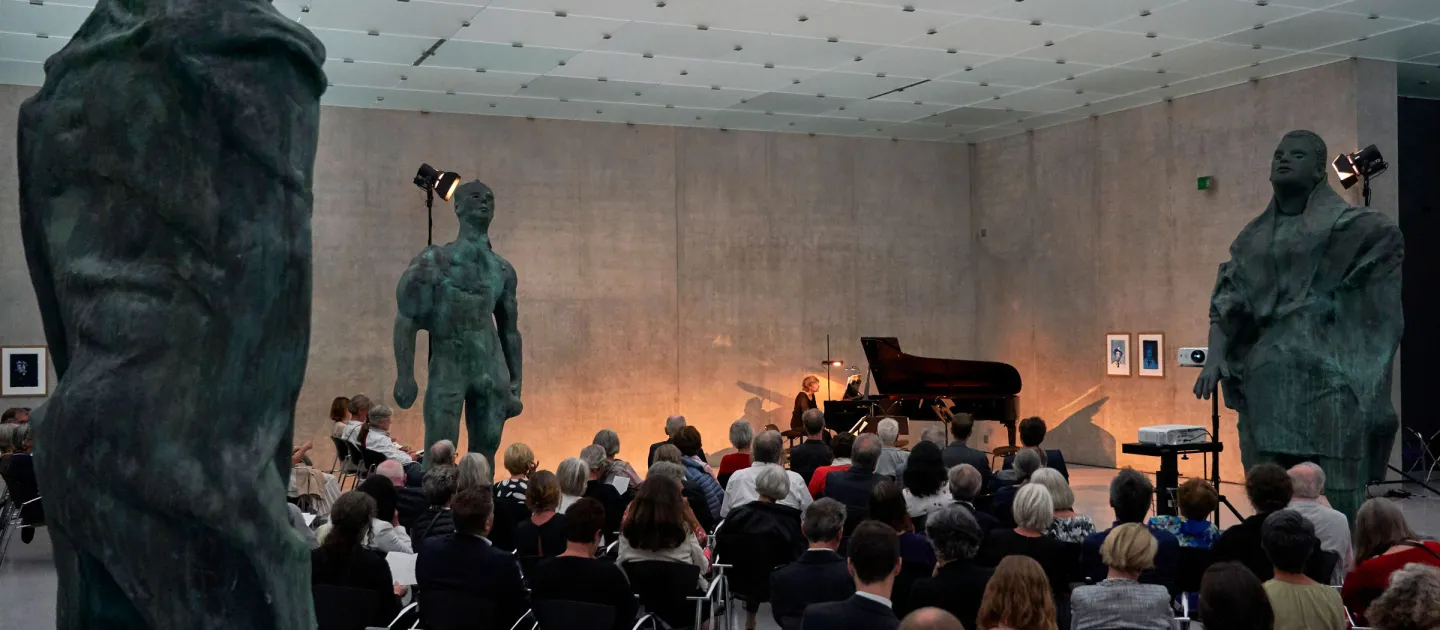 Szene aus "Konzert im KUB", 2019 im Kunsthaus Bregenz, Publikum bestaunt Dame am Klavier, drei übergroße Bronzefiguren umgeben das Publikum