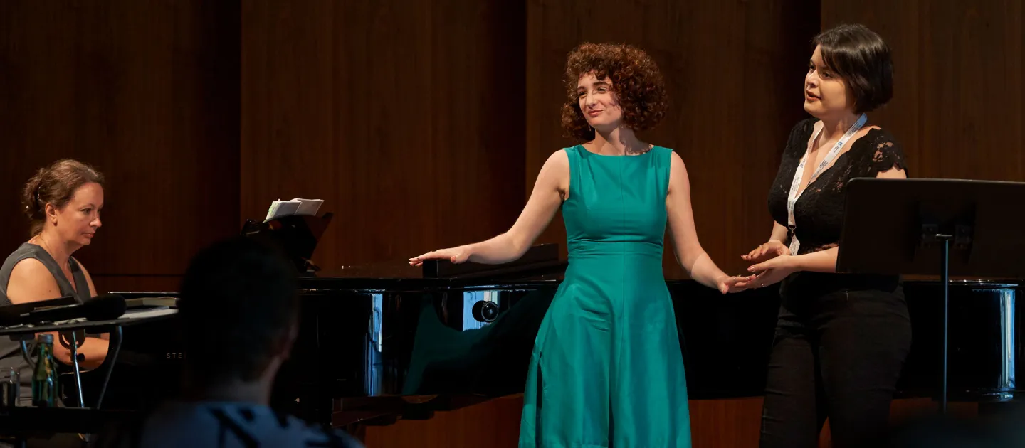 Szene aus "Meisterklasse Dmitry Vdovin", 2019 im Festspielhaus, drei Frauen auf der Bühne, eine im grünem Kleid und hält die Hand der danebenstehenden Frau in schwarzem Kleid, Pianistin am Flügel sitzend