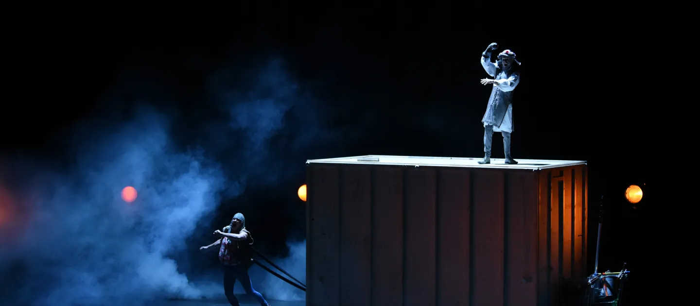 Szene aus "Don Quijote", 2019 im Festspielhaus, kostümierte Darsteller:in steht auf großem Kasten, zwei weitere kostümierte Darsteller unten am Kasten in ziehender Position befestigt