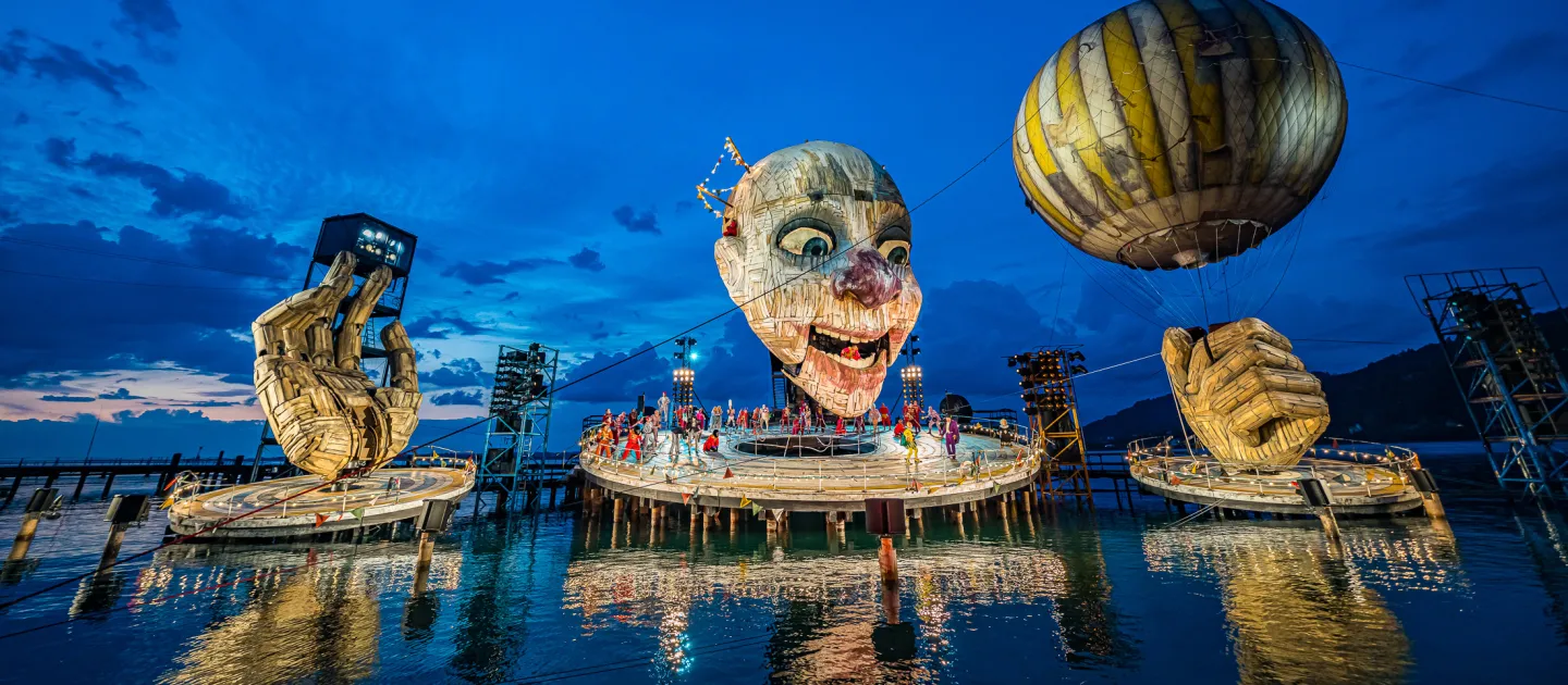 Szene aus "Rigoletto", 2019 auf der Seebühne, riesiger Kopf mit leicht geöffnetem Mund, zwei große Hände, eine Hand hält einen Ballon, mehrere kostümierte Darstellende auf der Bühnenplattform unter dem Kopf