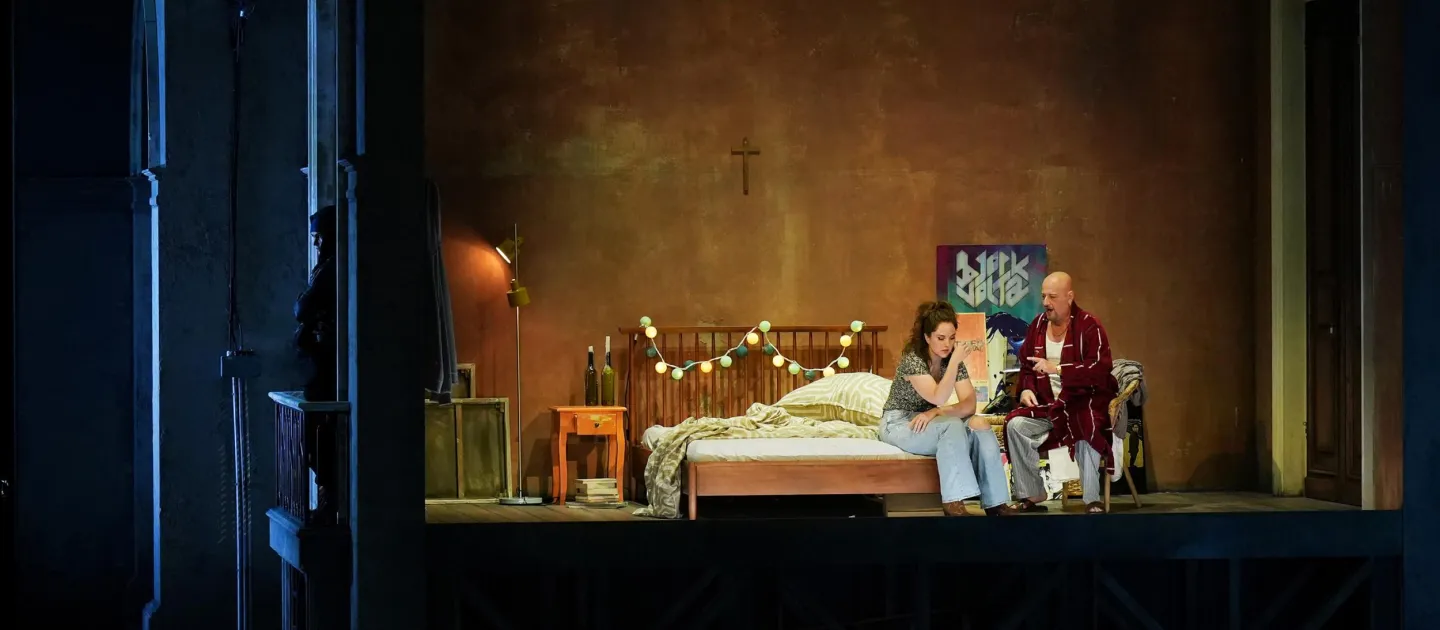 Szene aus "Tancredi", 2024 im Festspielhaus, zwei Personen auf einem Bett, eine Frau und ein Mann, im Hintergrund ein Kreuz an der Wand, Lichterkette über dem Bettende, Nachtkästchen und Lampe links neben dem Bett 