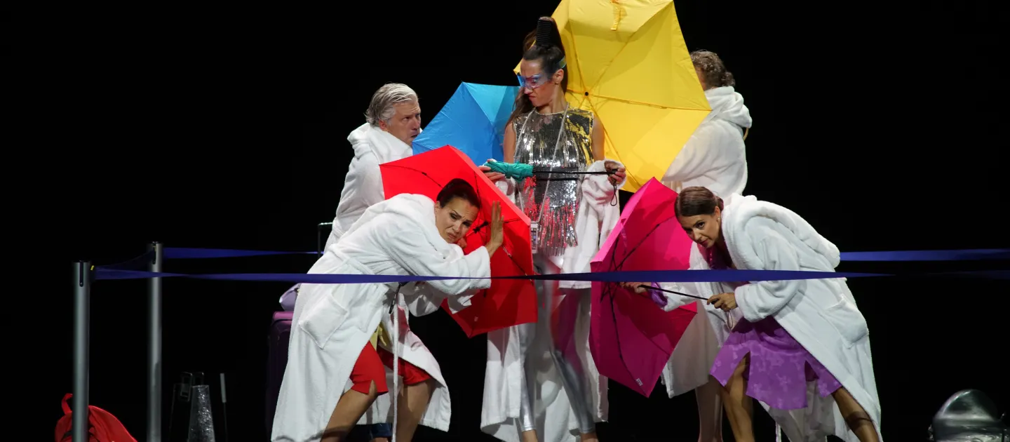 Szene aus "Impresario Dotcom", 2020 im Festspielhaus, fünf Personen, in der Mitte eine Darstellerin in glänzendem Kleid, die anderen tragen weiße Bademantel und halten bunte Regenschirme