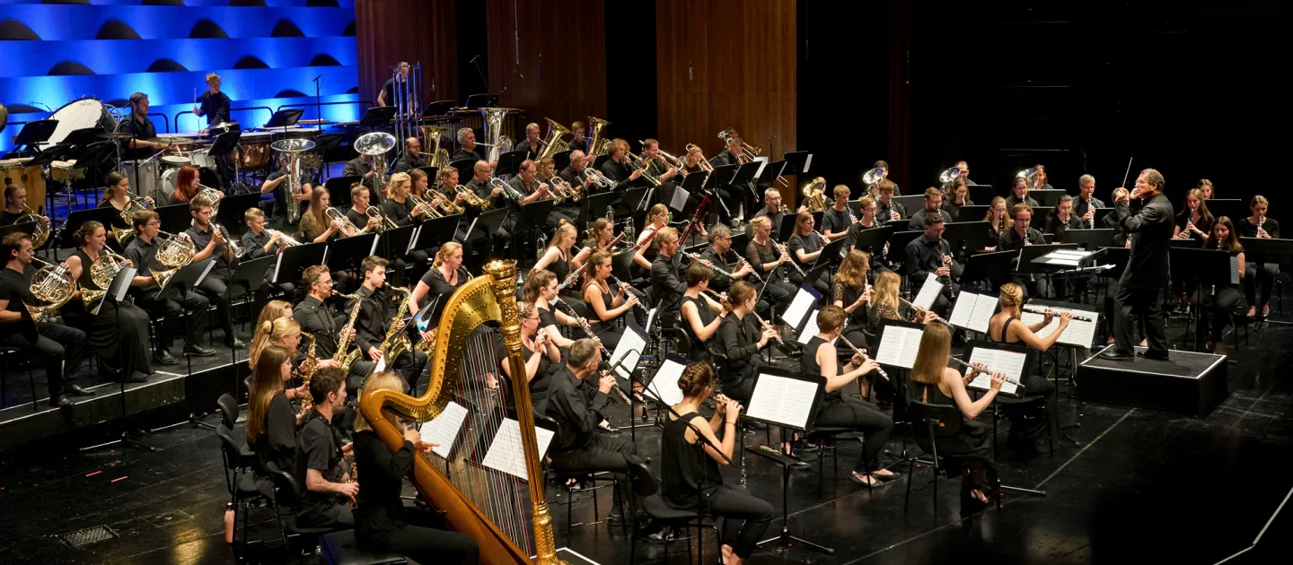 Szene aus "Brass Eroico", 2021 im Festspielhaus, großes spielendes Orchester mit Streichern, Bläsern und einer Harfe, Dirigent davorstehend