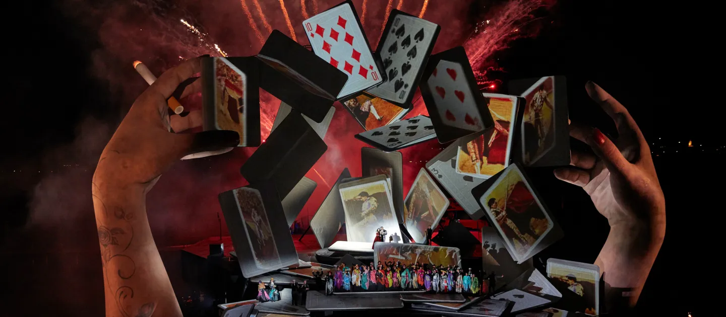 Szene aus "Carmen", 2018 auf der Seebühne, große Hände über dem Wasser, schwebende Spielkarten im Bogen, kostümierte Darstellende im Vordergrund auf der Bühnenfläche, rotes Feuerwerk im Hintergrund