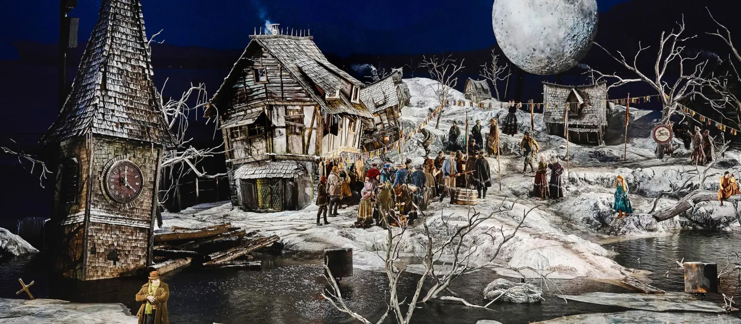 Szene aus "Der Freischütz", 2024 auf der Seebühne, schneebedecktes Dorf, links ein Turm mit Uhr, im Hintergrund ein großer Mond, viele kostümierte Darstellende auf der Bühne verteilt
