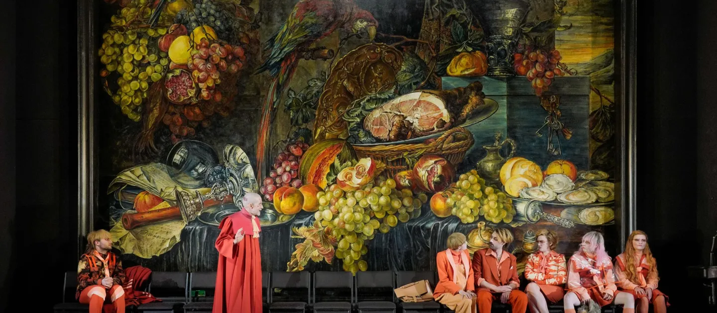 Szene aus "Der zerbrochene Krug", 2024 im Theater am Kornmarkt, sieben Darstellende in rot-orangen Kostümen vor einem großen Stillleben-Gemälde mit Früchten, Fleisch und einem Papagei