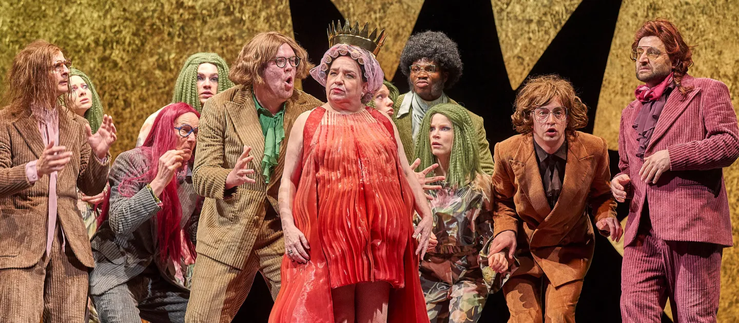 Szene aus "Die gefesselte Phantasie", 2023 im Festspielhaus, elf kostümierte Darstellende, zentral eine Person in rotem Kleid und mit Krone auf dem Kopf