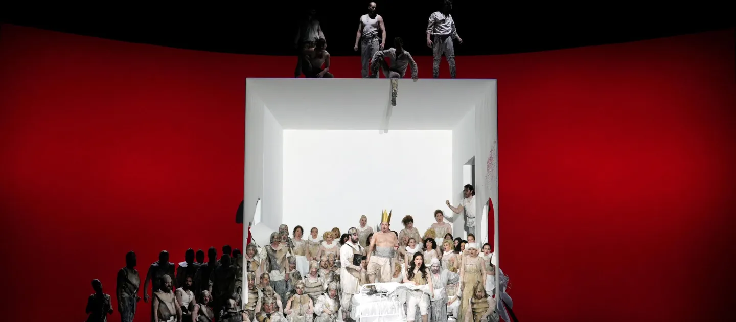 Szene aus "Ernani", 2023 im Festspielhaus, viele weiß kostümierte Darstellende in großer Kubusstruktur, roter Hintergrund, links weitere Darstellende im Schatten