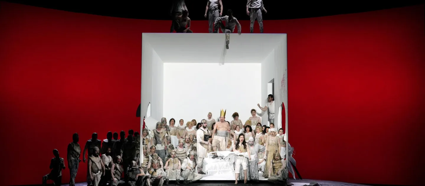 Szene aus "Ernani", 2023 im Festspielhaus, viele weiß kostümierte Darstellende in großer Kubusstruktur, roter Hintergrund, links weitere Darstellende im Schatten