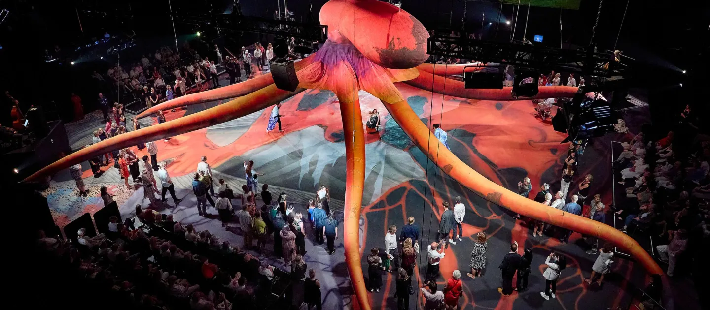 Szene aus "Hold Your Breath", 2024 im Festspielhaus, großer, roter Oktopus in der Mitte, umgeben von Zuschauende 