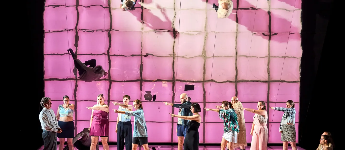 Szene aus "Die Judith von Shimoda", 2023 im Festspielhaus, zwölf kostümierte Darstellende, elf auf eine Person zeigend, rosa-farbender Boden, spiegelnde Wand im Hintergrund