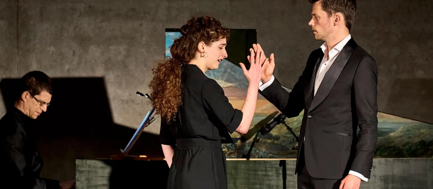 Szene aus "Konzert im KUB", 2023 im Kunsthaus Bregenz, drei Personen, zwei im Vordergrund an der rechten Hand haltend, eine im Hintergrund am Flügel