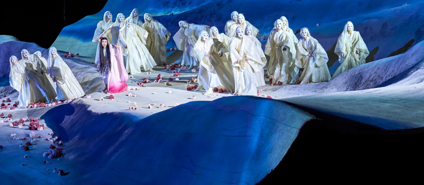 Szene aus "Madame Butterfly", 2022 auf der Seebühne, wellenförmige Bühne, rosafarbene Blüten auf dem Boden verteilt, viele Darstellende in weißen, langen Gewändern mit weißen Gesichtsmasken, links im Bild die Darstellende Madame Butterfly mit langem, dunklen Haar und rosafarbenem Gewand