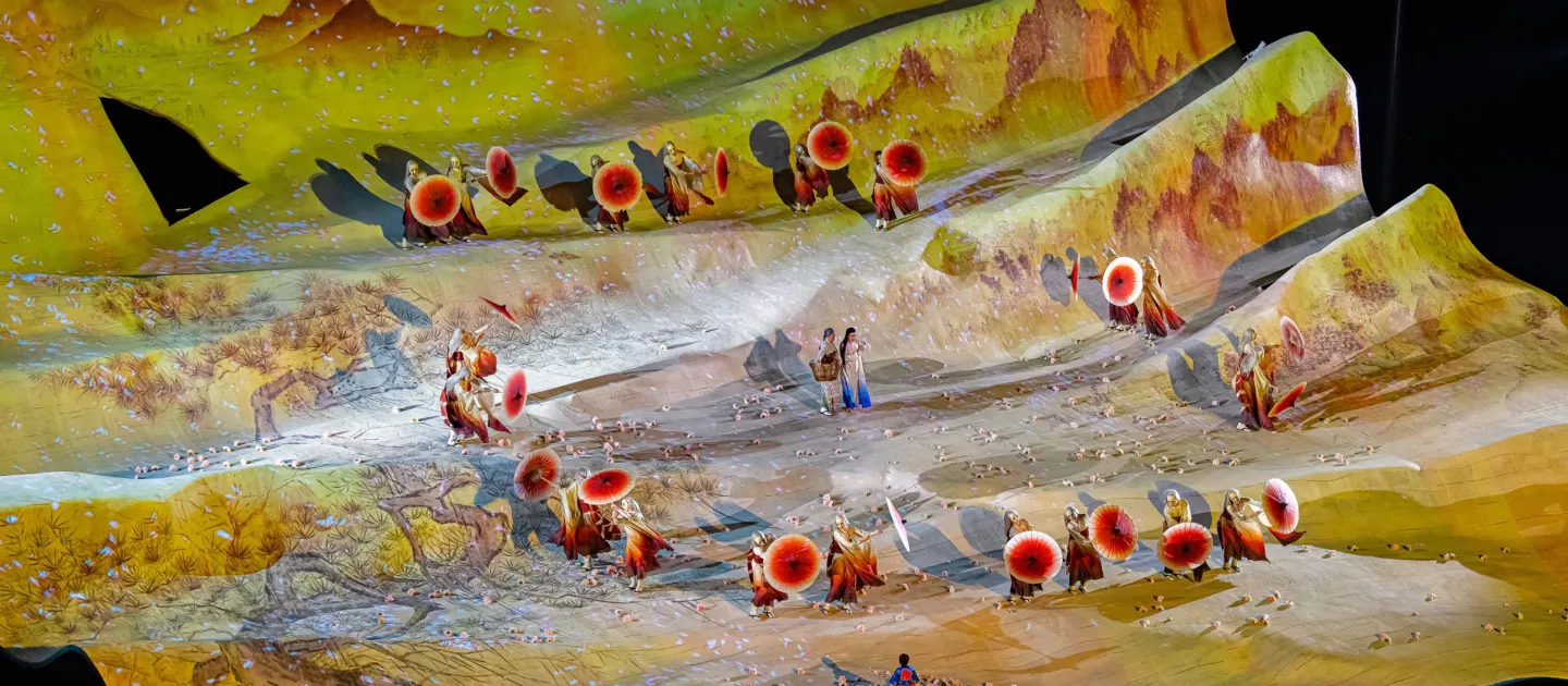 Szene aus "Madame Butterfly", 2023 auf der Seebühne, wellenförmige Bühne, bunte Projektion einer Berglandschaft mit Bäumen, Darstellende kreisförmig verteilt, zwei Darstellende zentriert