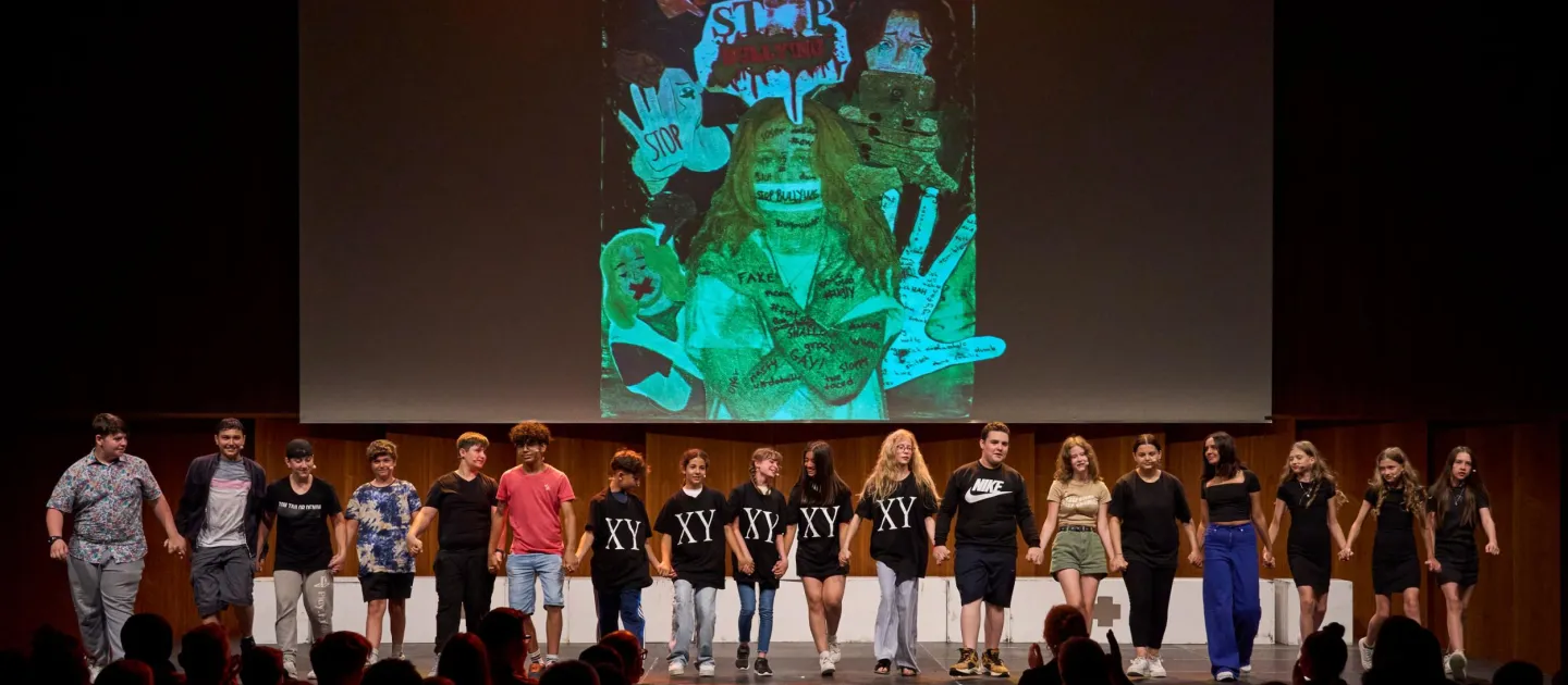 Szene aus "MINT-Projekt", 2023 im Festspielhaus, in einer Reihe stehende Jugendliche auf der Bühne, im Hintergrund eine Projektion mit Händen und Figuren in grüner Farbe