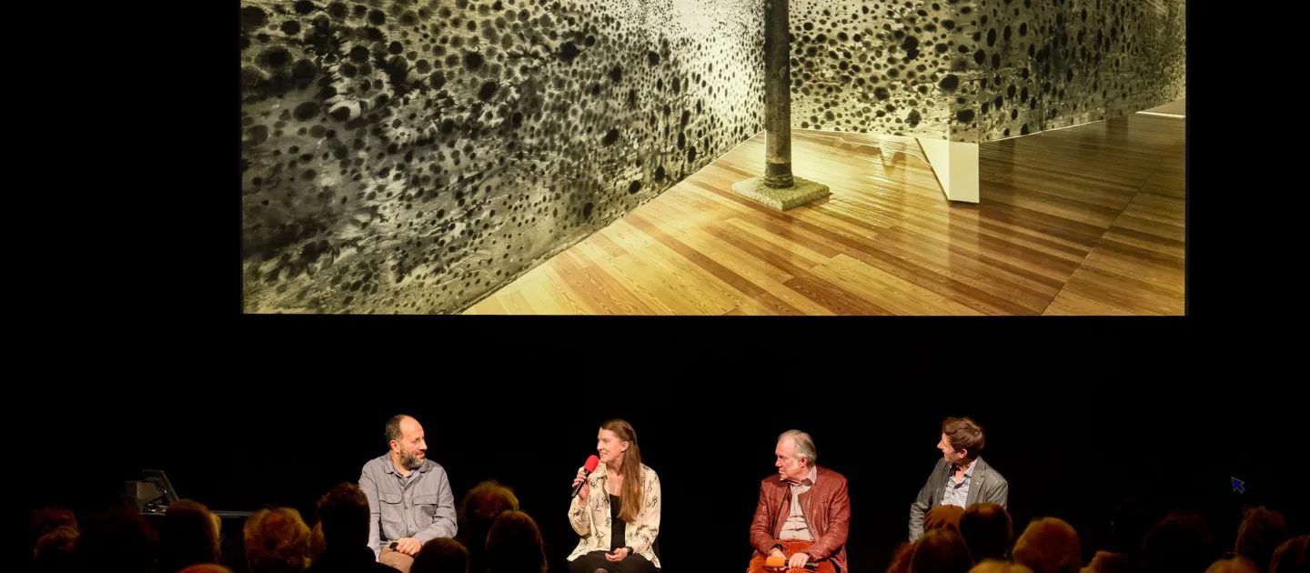 Szene aus "Einblick 2", 2023 im Festspielhaus, vier Personen auf der Bühne vor einem Publikum, im Hintergrund ein Bild eines großformatigen Kunstwerks mit schwarzen Punkten auf hellem Untergrund
