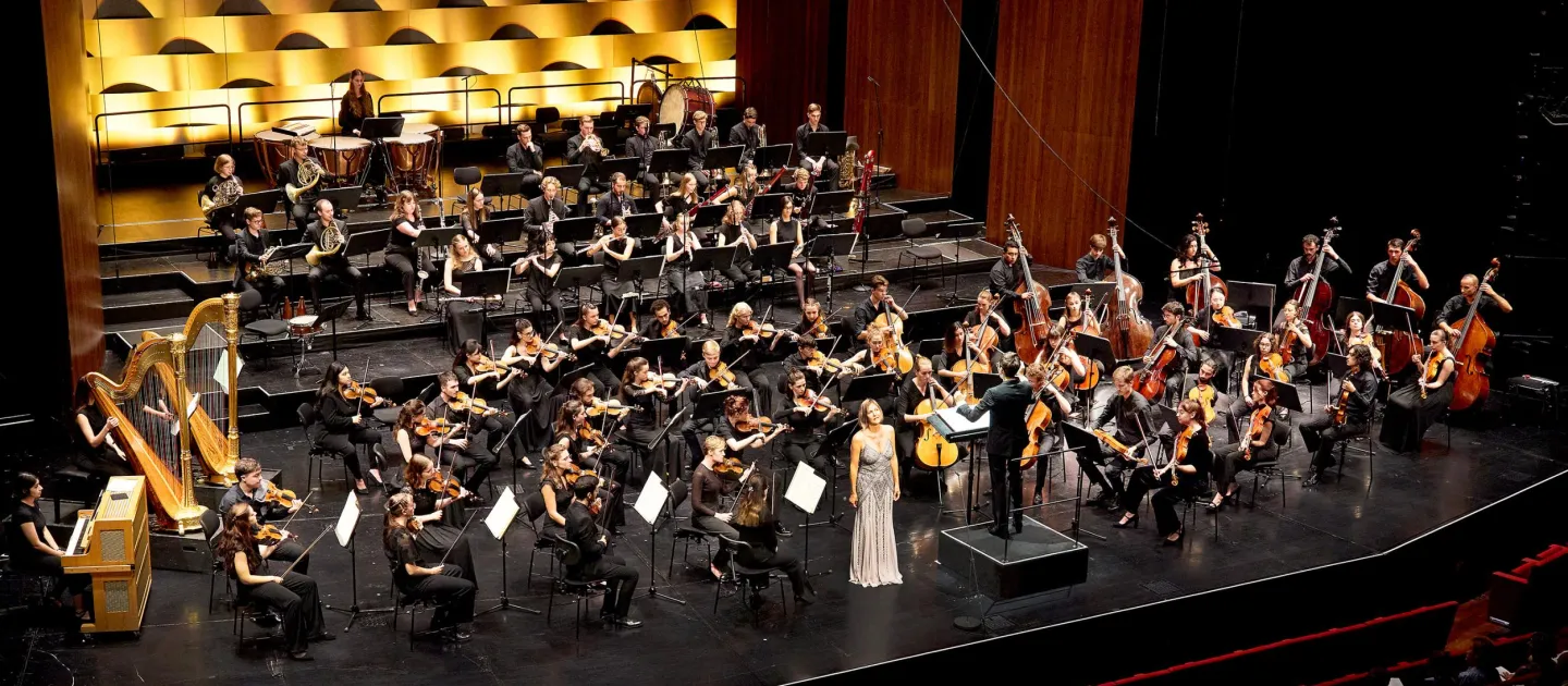 Szene aus "Orchesterakademie", 2024 im Festspielhaus, Orchester sitzend, Dirigent und Sängerin in hellem Kleid stehend