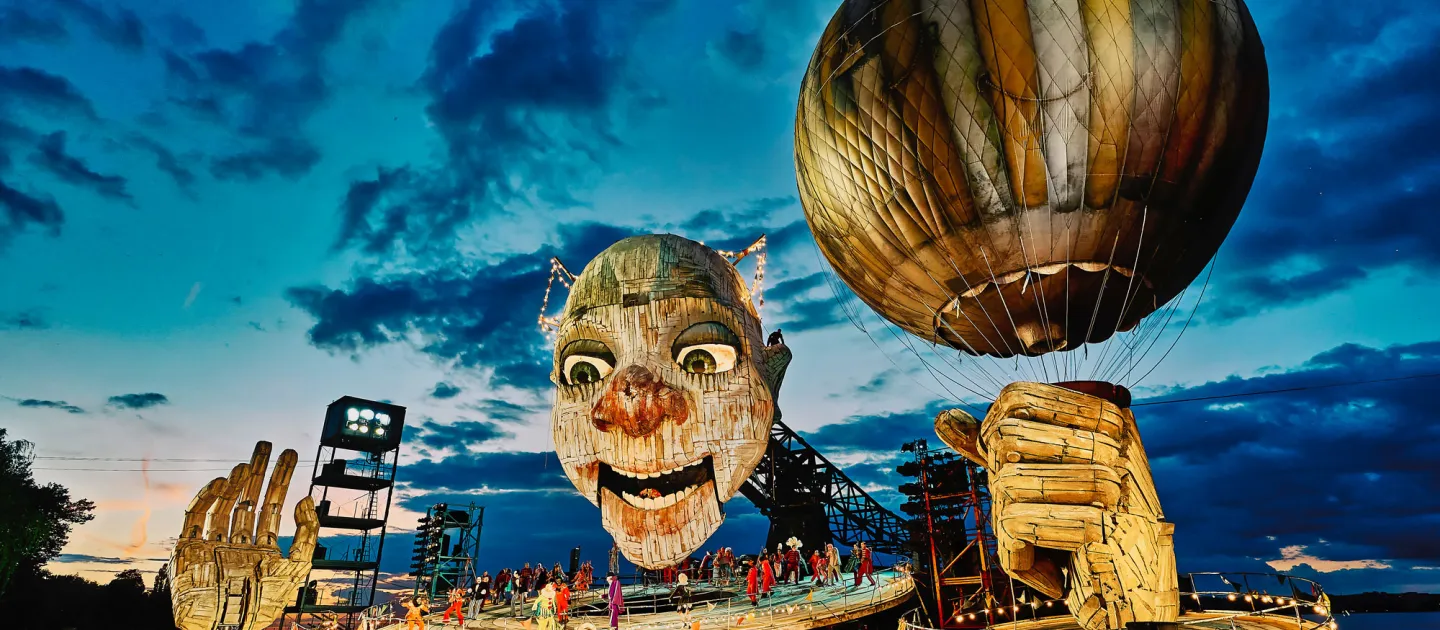 Szene aus "Rigoletto", 2021 auf der Seebühne, riesiger Kopf mit leicht geöffnetem Mund, zwei große Hände, eine Hand hält einen Ballon, mehrere kostümierte Darstellende auf der Bühnenplattform unter dem Kopf
