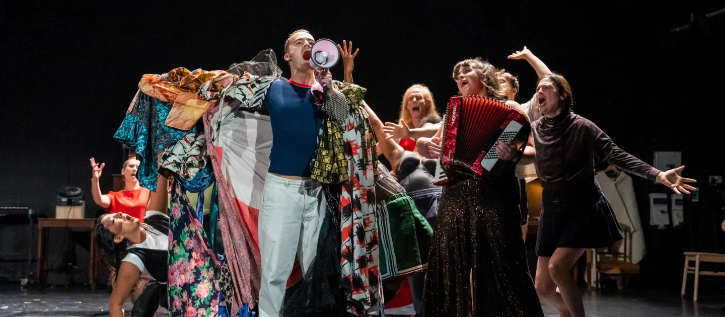 Szene aus "The Faggots and Their Friends Between Revolutions", 2023 im Festspielhaus, acht singende und kostümierte Darstellende, eine Person mit Megafon in der Hand, eine weitere mit einem Akkordeon