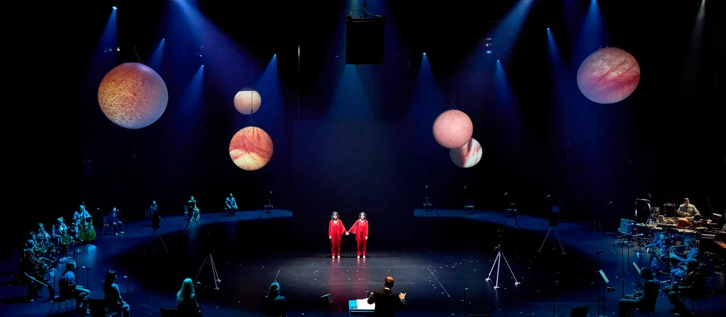 Szene aus "Unmögliche Verbindung", 2024 im Festspielhaus, von der Decke hängende Planetenmodelle, in der Mitte zwei sich an Händen haltende Darstellende in rotem Kostüm