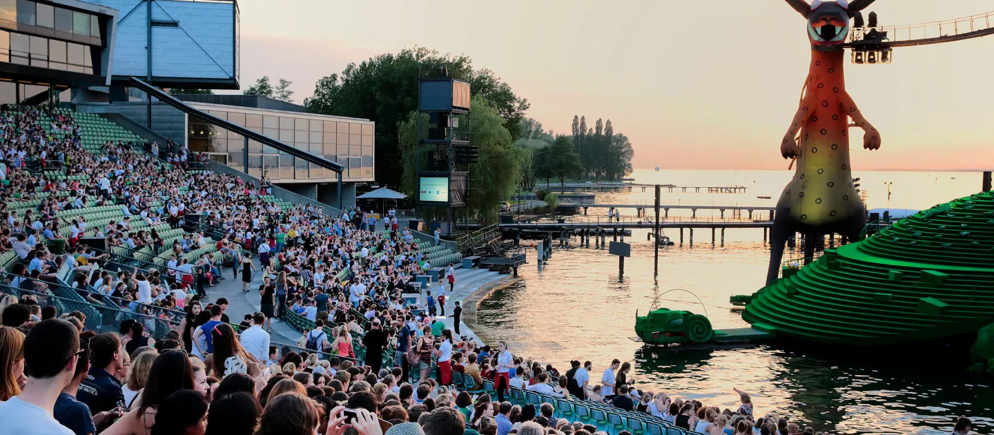 Szene aus "crossculture", 2014 an diversen Spielorten, viele Menschen sitzen auf der Tribüne, rechts ein Teil des Bühnenbilds von "Die Zauberflöte", im Hintergrund der Sonnenuntergang