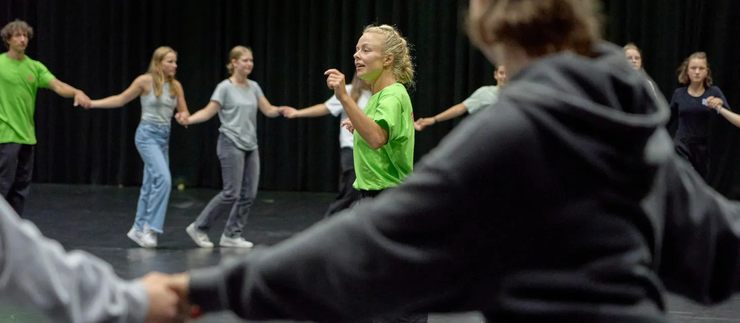 Szene aus "Young People's Night", 2024, eine Gruppe junger Menschen in einem Kreis, sich an den Händen haltend und tanzend vor einem schwarzen Hintergrund