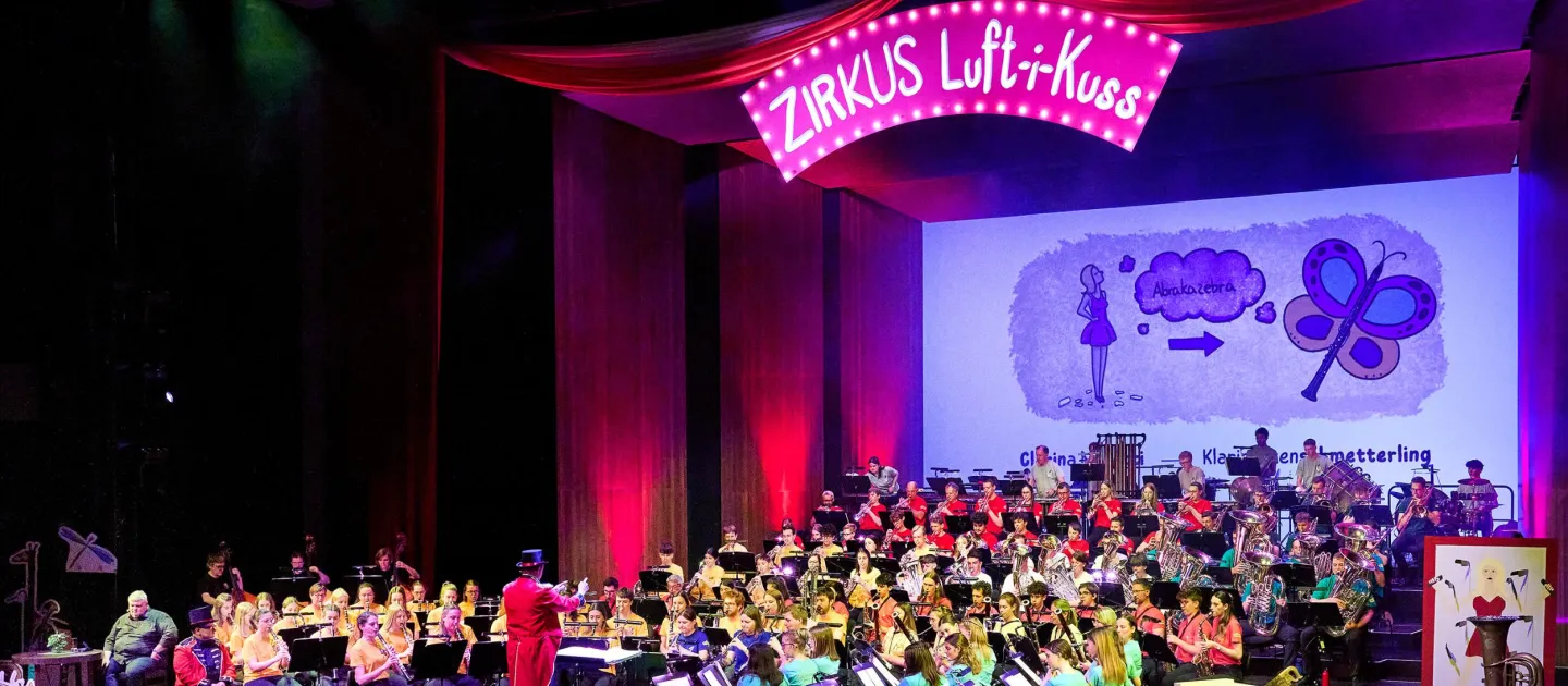 Szene aus "Zirkus Luft-i-Kuss", 2024 im Festspielhaus, Orchester in bunter Kleidung, Dirigent in rotem Frack, Banner mit Schriftzug "ZIRKUS Luft-i-Kuss" an der Decke