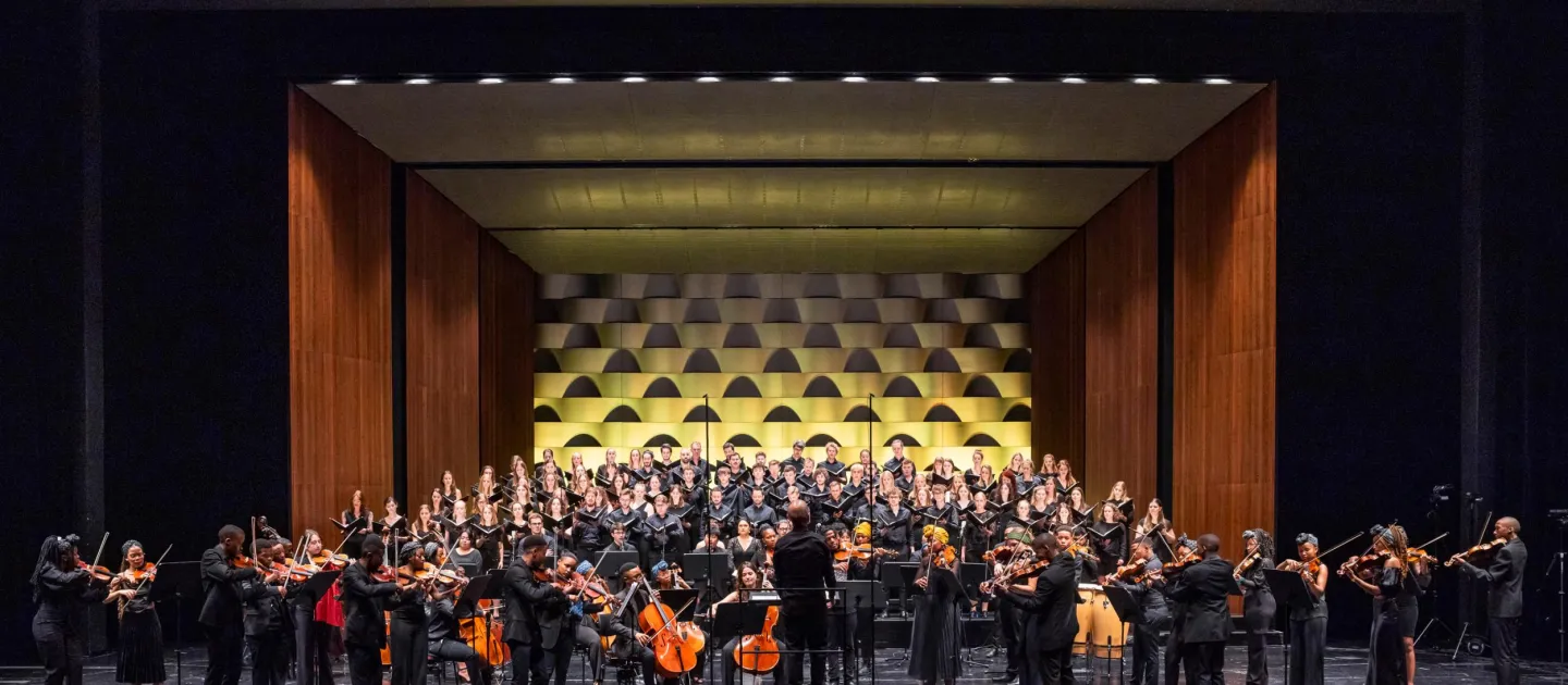 Szene aus "Zwischen Himmel und Erde", 2023 im Festspielhaus, Orchester stehend und spielend im Vordergrund auf der Bühne, Chor im Hintergrund