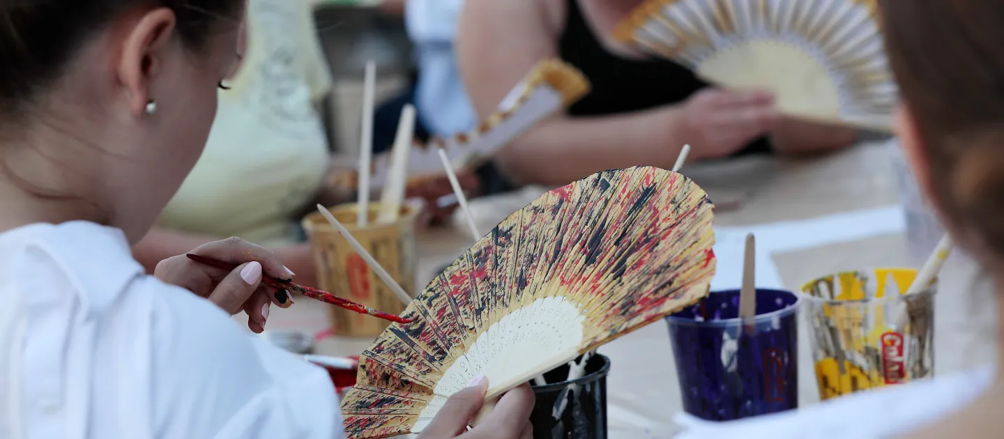 Szene aus "crossculture", 2018 an diversen Spielorten, Detailaufnahme, Person bemalt einen Fächer aus Holz mit Pinsel und Farbe