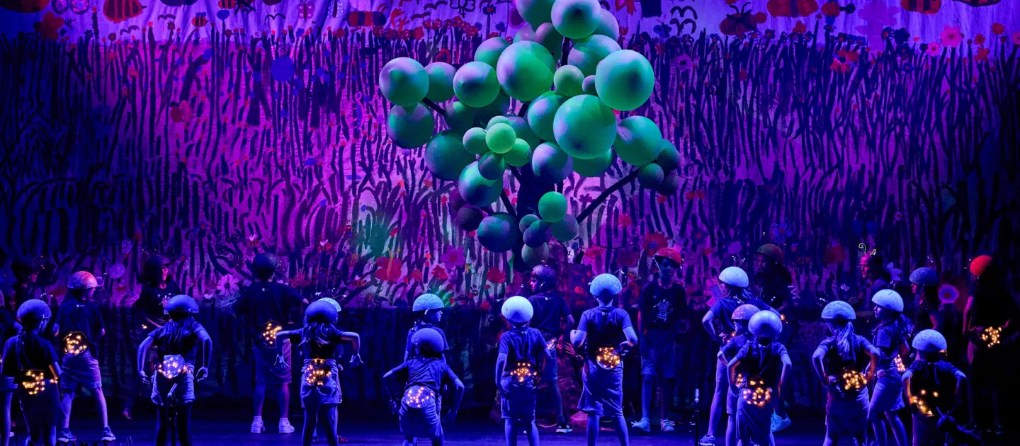 Szene aus "Junge Festspiele", 2022 im Festspielhaus, viele Kinder mit Rücken zum Publikum und Helmen auf dem Kopf, grüne Luftballons, im Hintergrund gemalte Wiese und Insekten, in Schwarzlicht