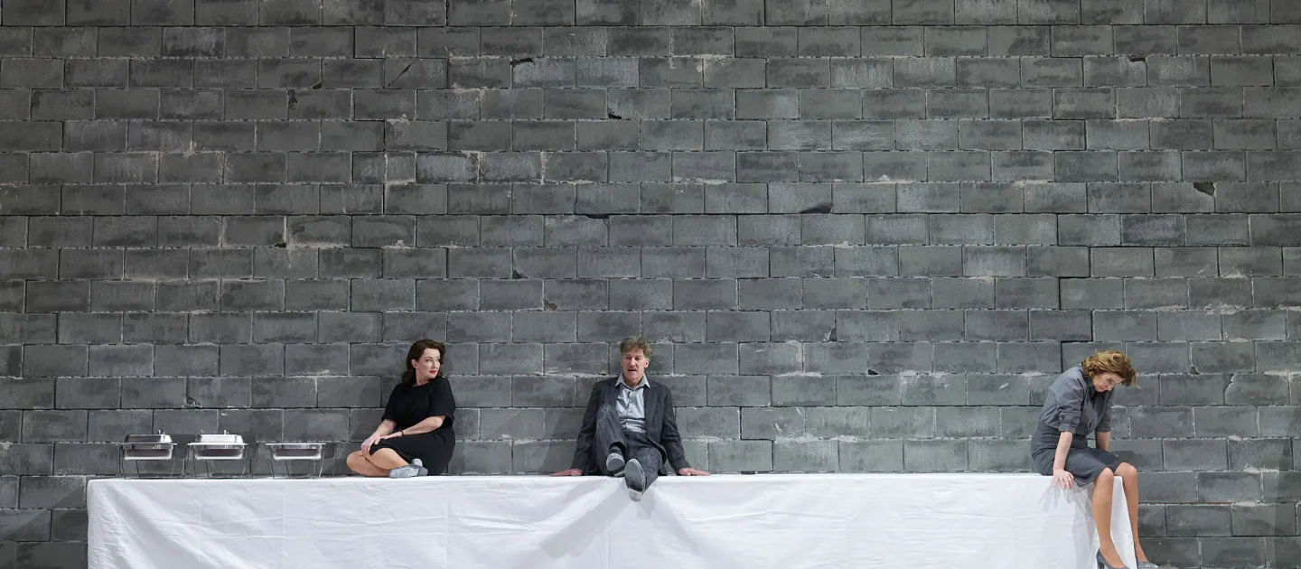 Szene aus "Geschlossene Gesellschaft", 2022 im Festspielhaus, drei kostümierte Darstellende, sitzend auf einer Tafel, Tisch mit weißer Tischdecke bis zum Boden bedeckt, graue Steinmauer als Hintergrund 